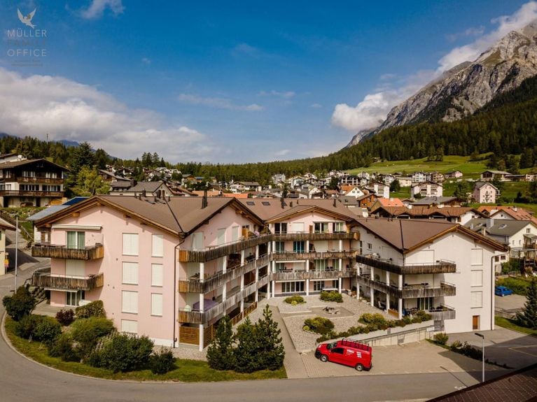 A multi-story building is surrounded by landscaped greenery, with several balconies visible on the exterior. The structure's architecture provides a blend of modern and traditional elements. A red vehicle is parked in the front area, with mountains rising in the background and a picturesque village layout.