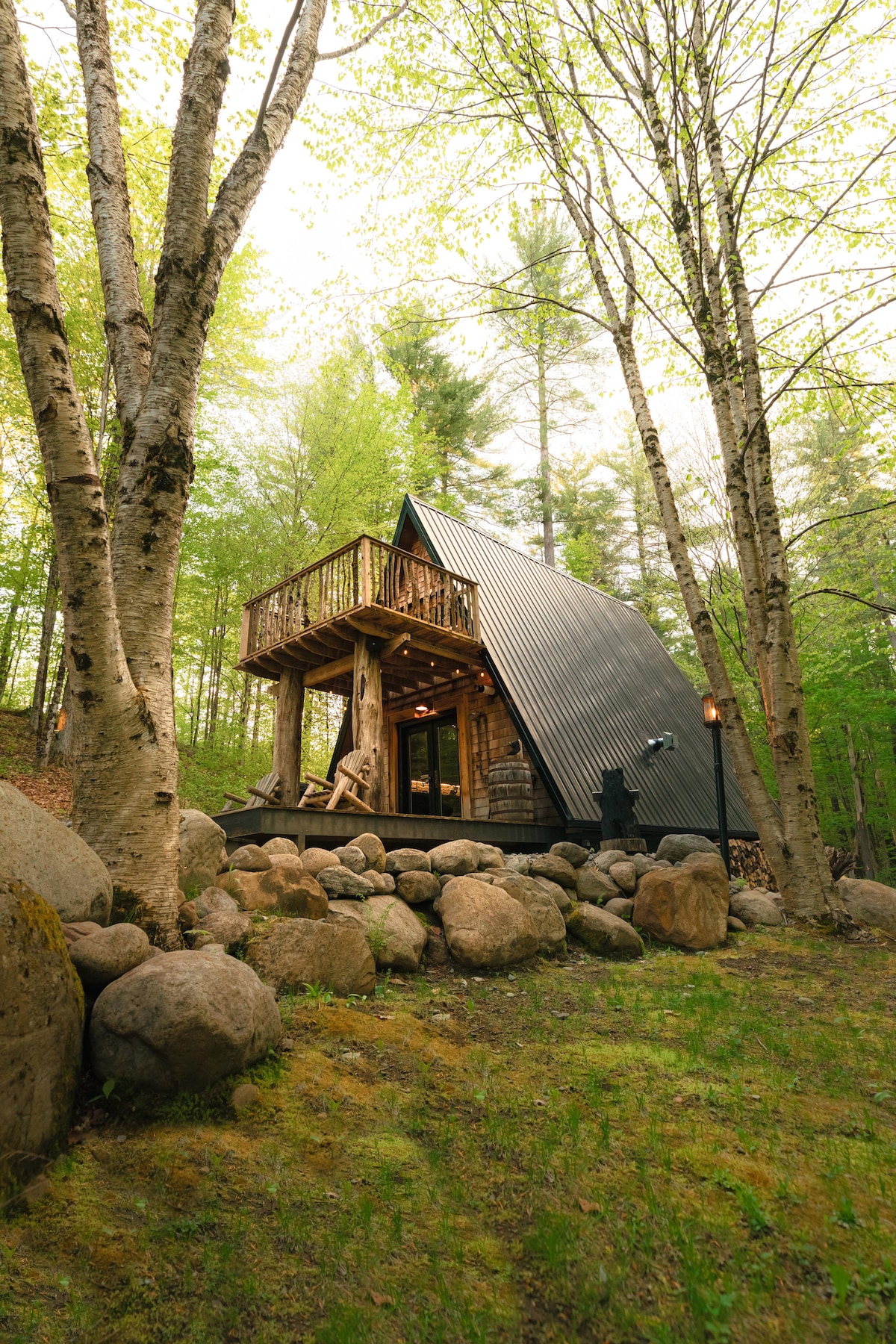 The A-frame cabin is surrounded by lush greenery, showcasing its distinctive steeply angled roof. Natural stone pathways lead to the entrance, where a cozy deck with rustic wooden seating overlooks the tranquil woodland setting.
