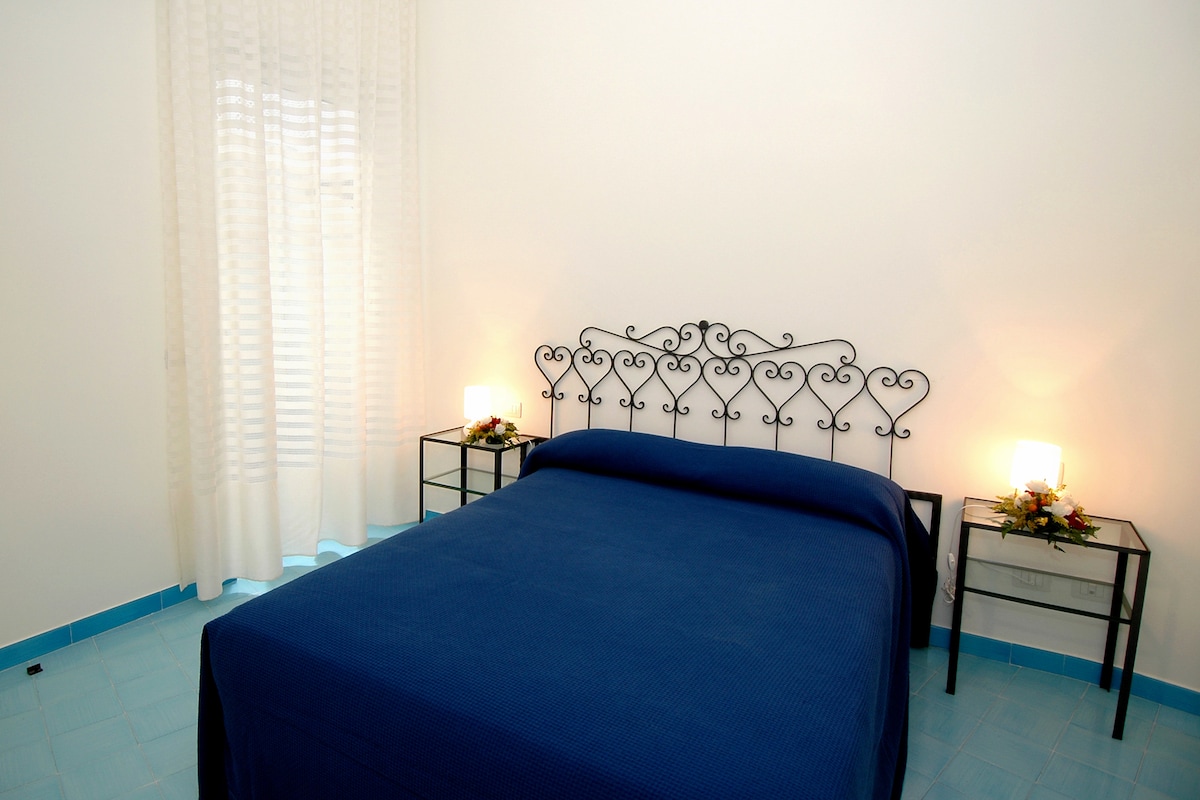 The sleeping area features a dark blue bedspread covering a spacious bed, with a decorative wrought-iron headboard. Two bedside tables with lamps flank the bed, providing soft lighting. Sheer white curtains allow natural light to enter, complementing the light blue tiled floor.