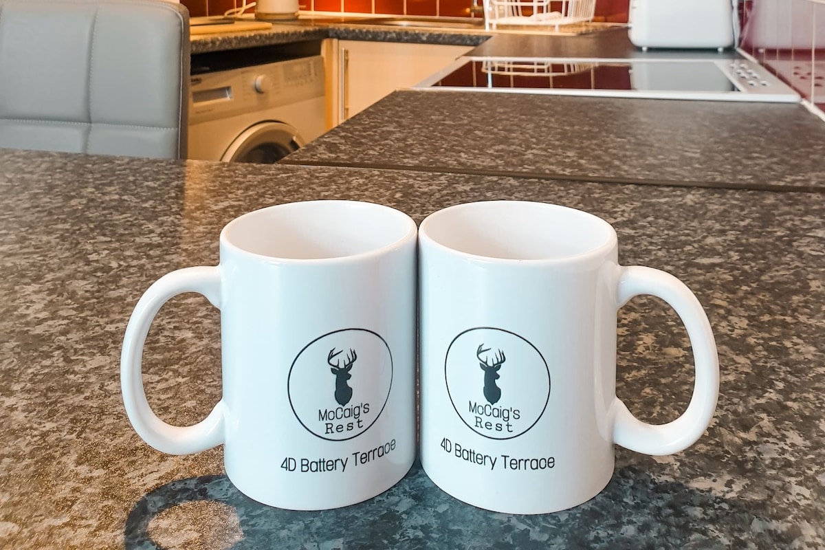Two white coffee mugs with a logo featuring a deer are placed on a granite countertop. The background reveals a modern kitchen environment, including a washing machine and cooking appliances within an open layout.