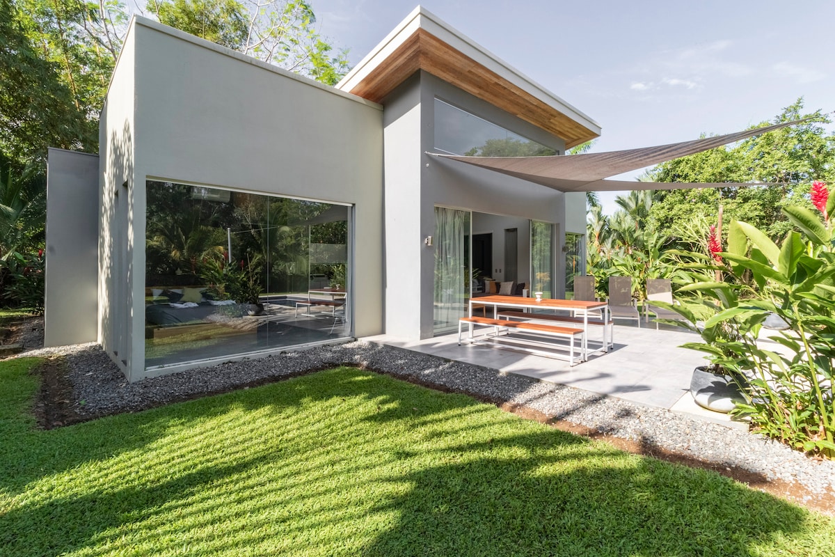 A modern exterior view of a house is shown, featuring large glass windows and a covered outdoor dining area. Lush greenery surrounds the property, with well-maintained grass and tropical plants visible. The architectural design emphasizes a blend of natural light and open space.