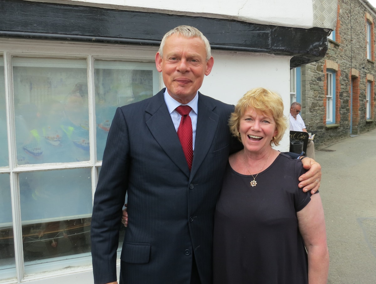 Two individuals are standing together outside a building with a white exterior and large windows. The setting appears to be in a village with stone and painted walls visible in the background. The people are dressed formally, with one wearing a suit and tie.