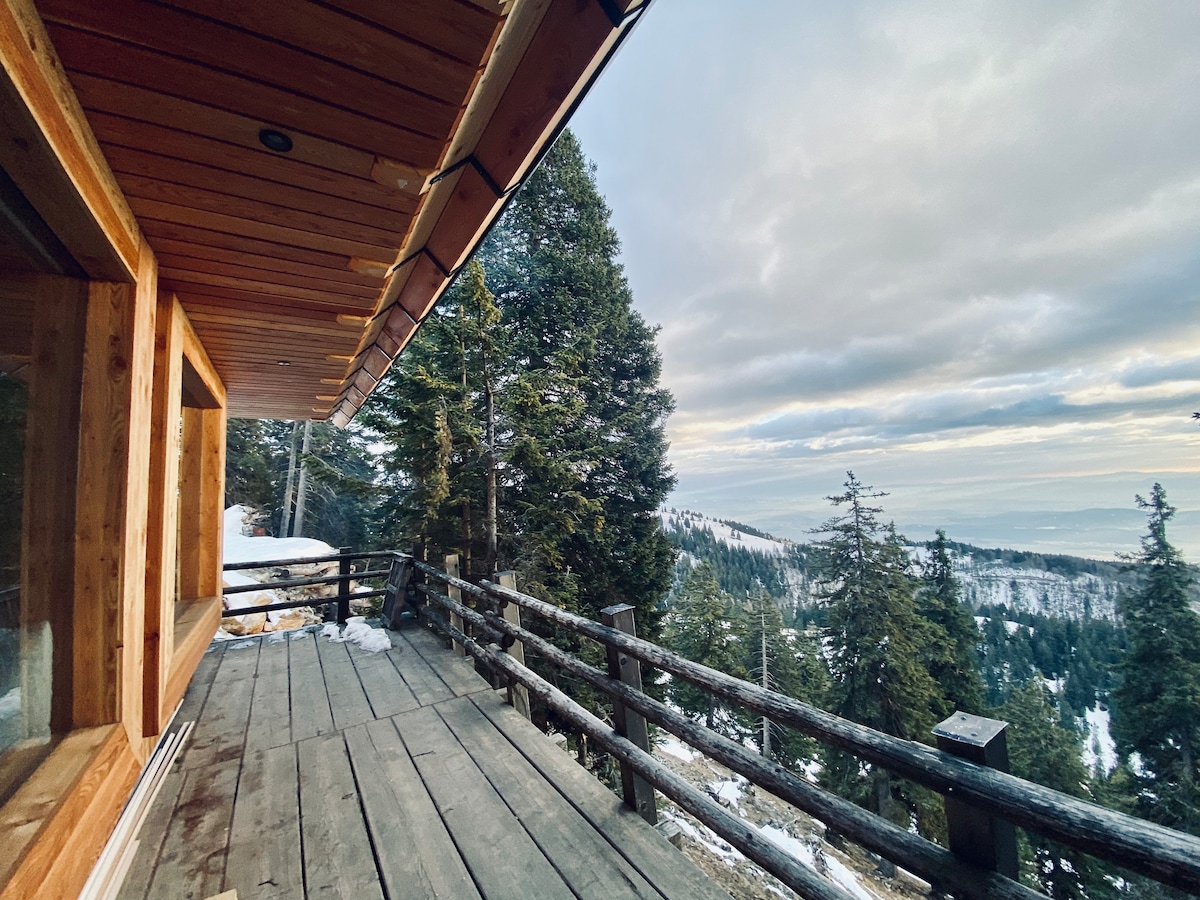 A wooden deck extends from the cabin, offering views of distant mountains and forested terrain. Snow covers the ground, and tall trees frame the scene. The cabin features large windows that bring natural light into the interior.