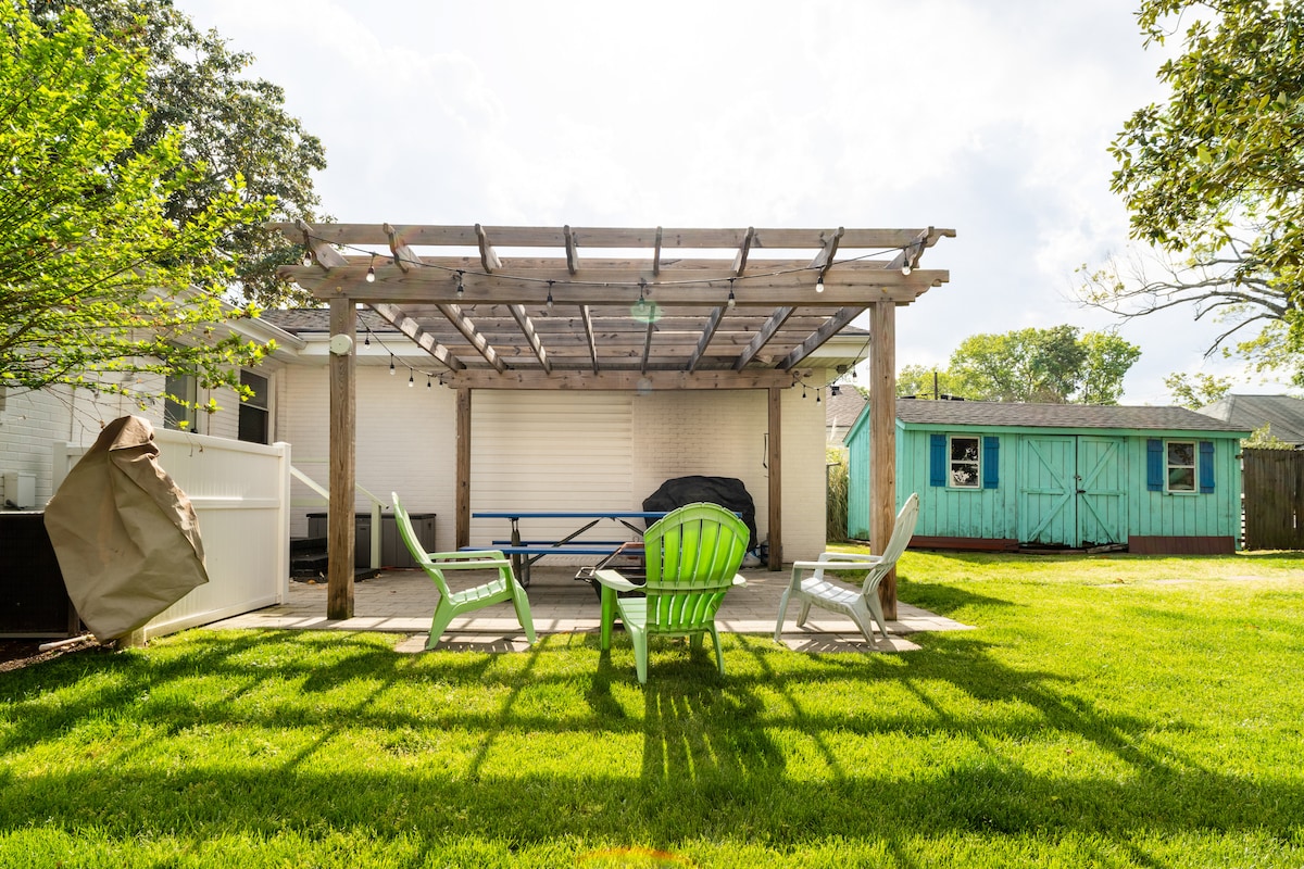 A serene backyard space features a wooden pergola overhead, providing shade and a relaxing seating area. Brightly colored adirondack chairs encircle a stone patio, with a well-maintained lawn extending beyond. A nearby shed adds a touch of charm to the outdoor setting.
