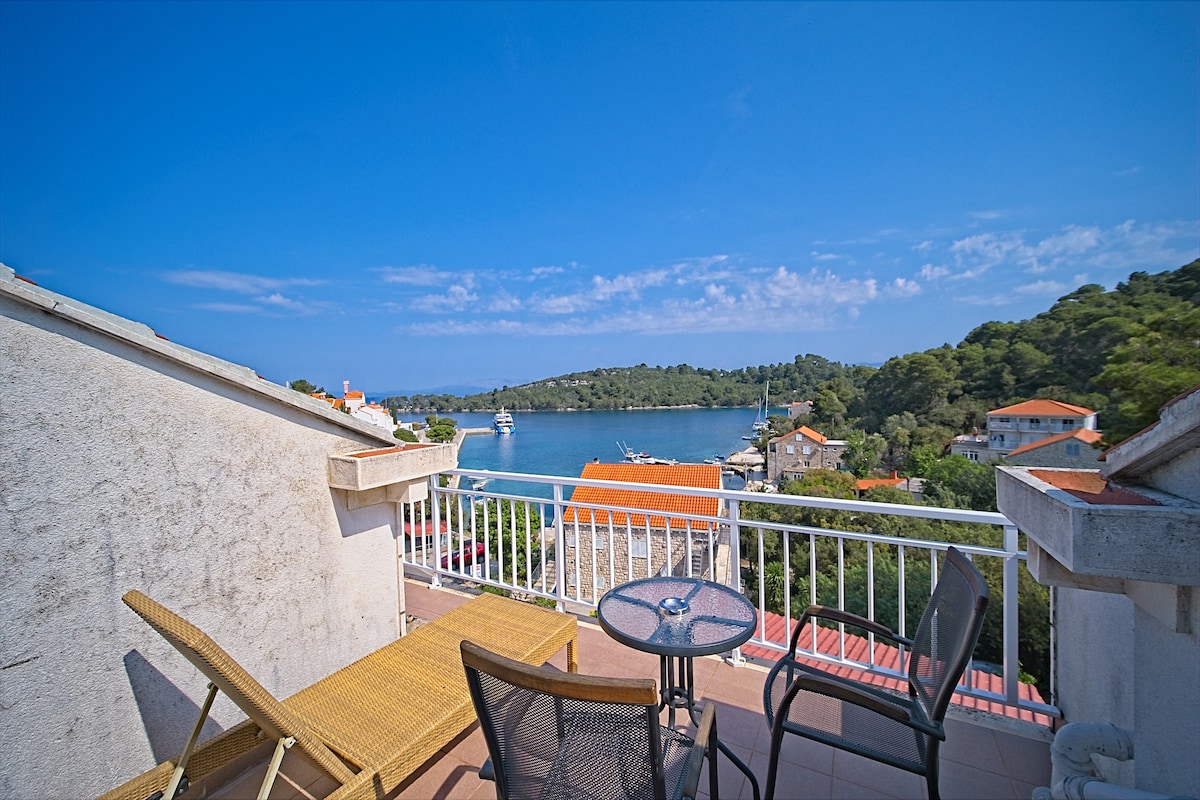 A private balcony is shown, furnished with a table and two chairs. The view captures a serene bay surrounded by lush greenery and scattered residential buildings. A clear blue sky enhances the coastal scenery, inviting relaxation and tranquility.
