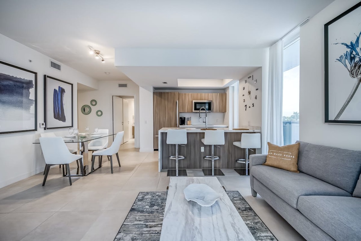 An open-concept living area features a gray sofa, a dining table for four, and a modern kitchen with wood cabinetry. Decorative artwork adorns the walls, and large windows allow natural light to fill the space, enhancing its welcoming atmosphere.