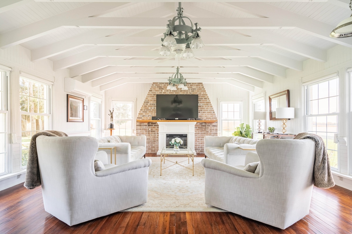A light-filled great room features a high ceiling with exposed beams, complementing the brick fireplace as a focal point. Two soft armchairs face a modern television, surrounded by large windows offering views of the outdoors. A coffee table sits on a plush area rug, enhancing comfort.