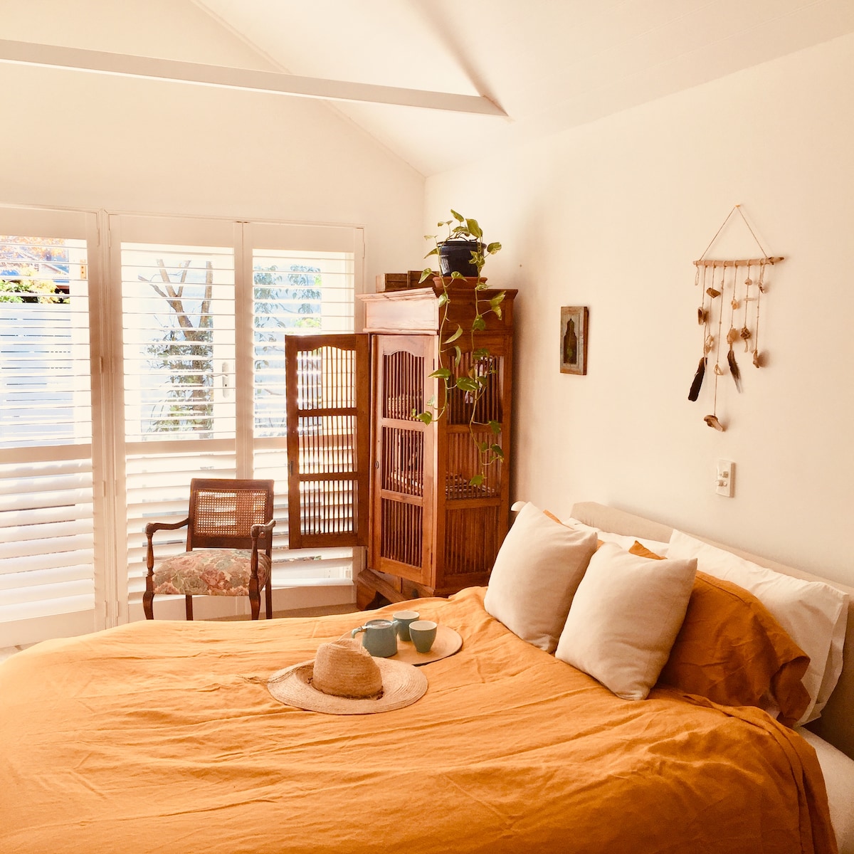 A comfortable bed with an orange duvet is positioned against a light wall. A wooden cabinet sits nearby, accompanied by a decorative chair. Natural light filters through shuttered windows, illuminating potted plants, while a simple wall hanging adds a touch of artistry.