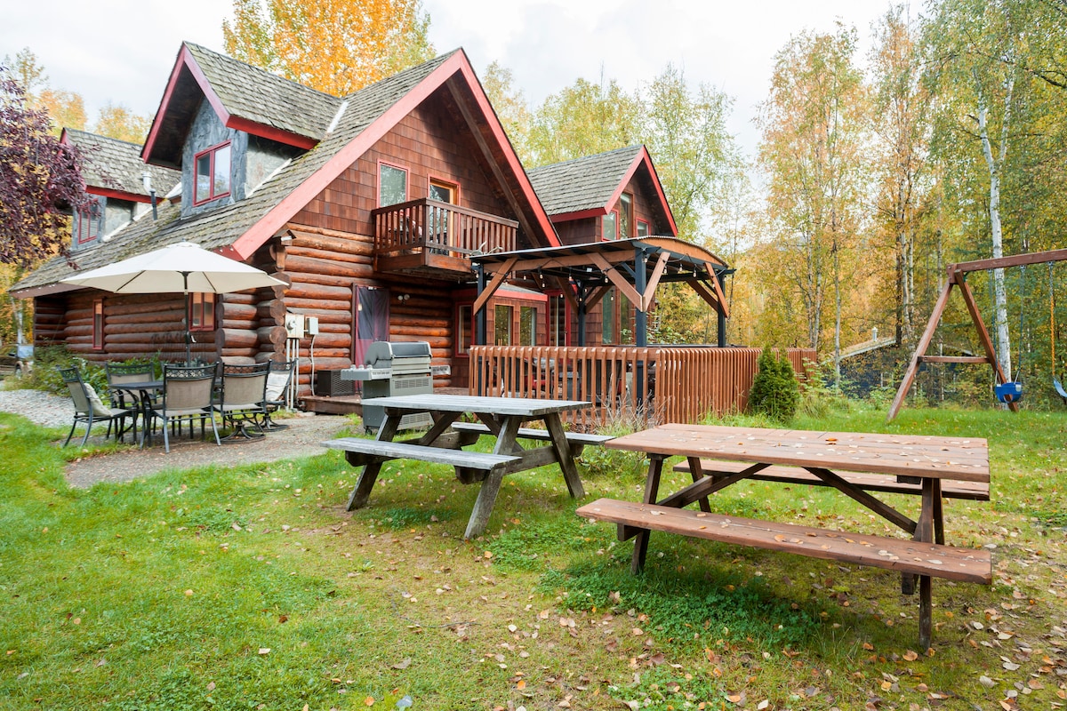 The exterior of a rustic log cabin is surrounded by vibrant fall foliage. Picnic tables are positioned on a grassy area, alongside a patio with outdoor seating under a large umbrella. A swing set is visible in the background, completing the inviting outdoor space.