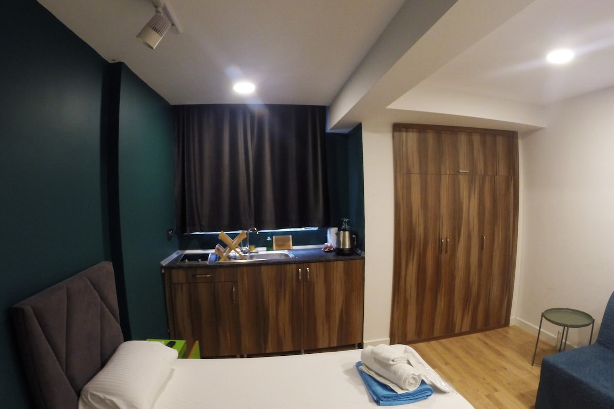 A contemporary studio apartment features a spacious bed with neatly arranged towels. A kitchenette area is visible, equipped with essential appliances and utensils. Dark curtains frame the window, while a wooden closet provides storage. Soft lighting enhances the modern design.