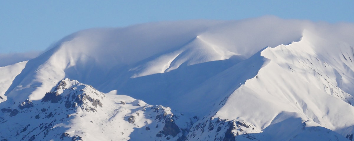 Snow-covered mountain peaks rise under a clear blue sky, with soft clouds clinging to the summit. The rugged terrain is visible, showcasing the natural beauty of the alpine landscape.