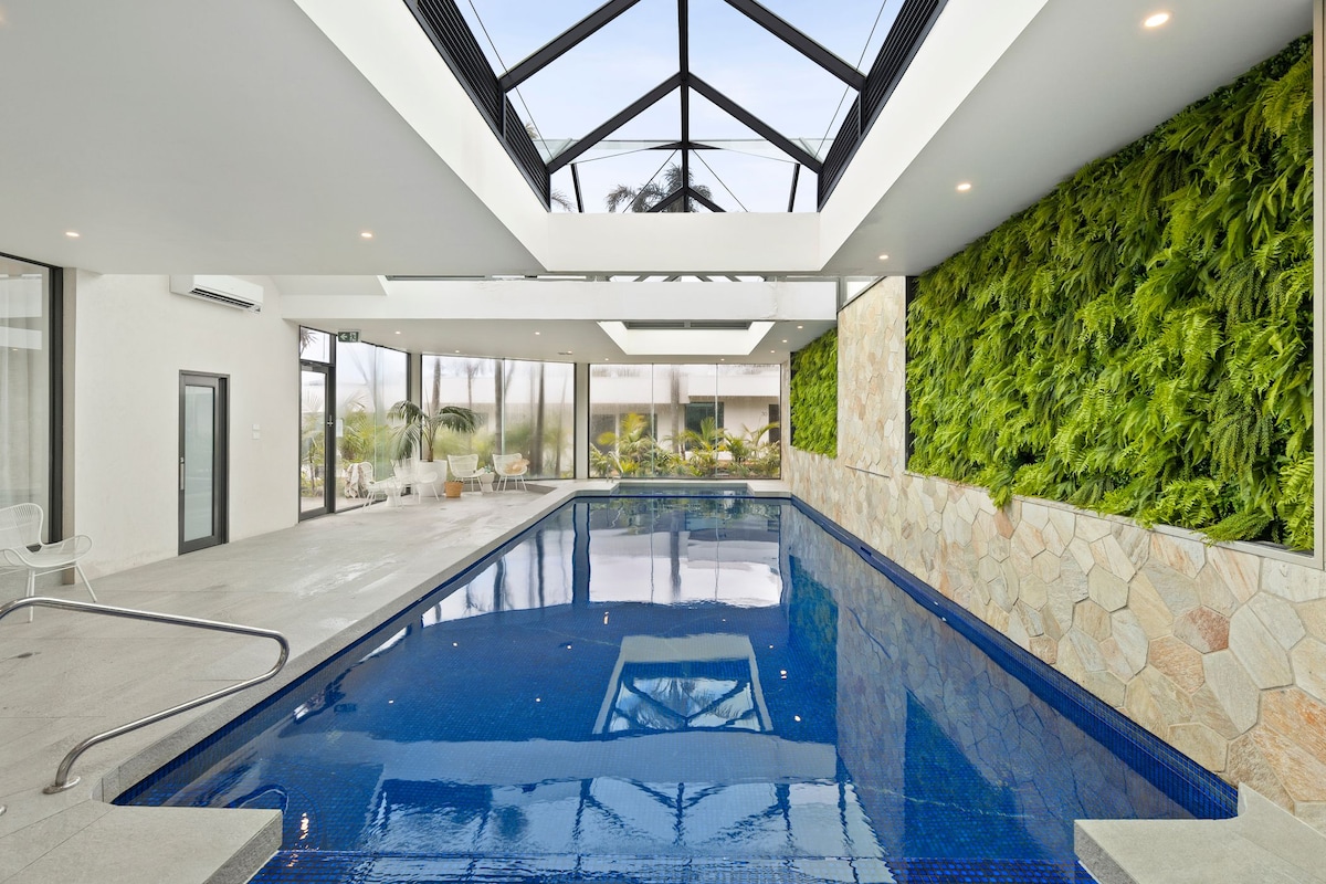 An indoor heated pool is showcased under a glass roof, providing natural light and a spacious ambiance. The water reflects the surroundings, complemented by a vibrant green wall of plants and natural stone accents on adjacent walls.