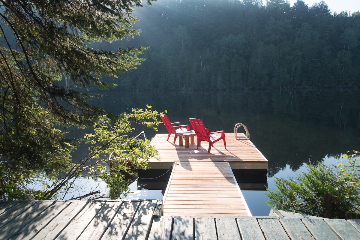 A wooden dock extends into calm waters, offering two red Adirondack chairs positioned around a small table. Lush greenery frames the scene, while distant tree-covered hills are reflected in the tranquil lake.