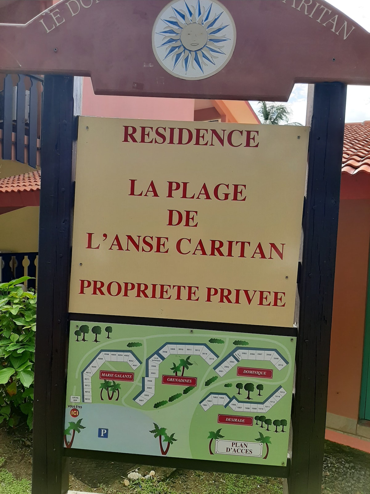A welcoming sign for the private residence, emblazoned with a sun emblem at the top. The name 'LA PLAGE DE L’ANSE CARITAN' is prominently displayed in bold red letters. Below, a detailed map of the property layout indicates various areas and facilities.
