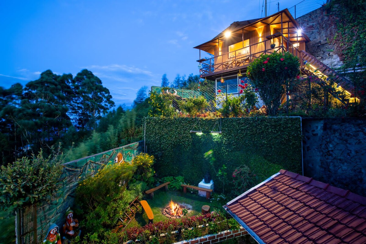 A hillside villa is captured in the twilight, showcasing a beautifully landscaped garden with vibrant greenery and a welcoming fire pit area. The villa features a spacious balcony and is set against a backdrop of misty hills, emanating a serene and inviting ambiance.