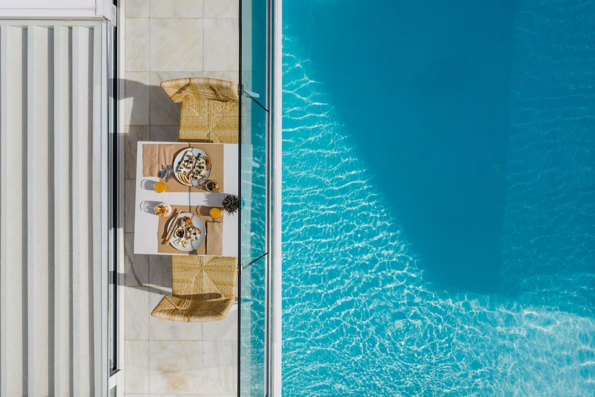 An aerial view captures a dining table set beside a sparkling pool. The table is adorned with plates and glasses, while two woven chairs provide seating. The pool's clear blue water reflects sunlight, enhancing the serene atmosphere of the outdoor space.