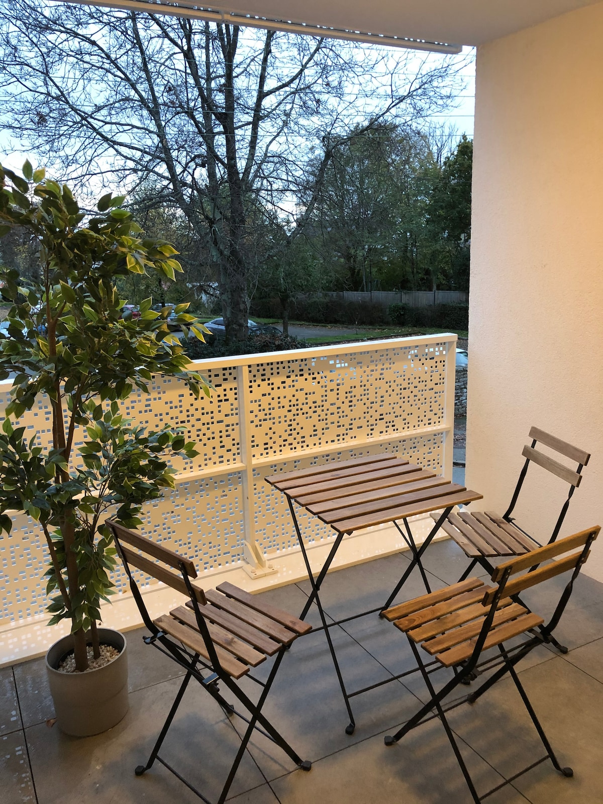 A small terrace features a modern metal balustrade with a perforated design. Two wooden foldable chairs and a matching table are arranged on the tiled floor. A potted plant adds a touch of greenery, offering a view of the surrounding trees.