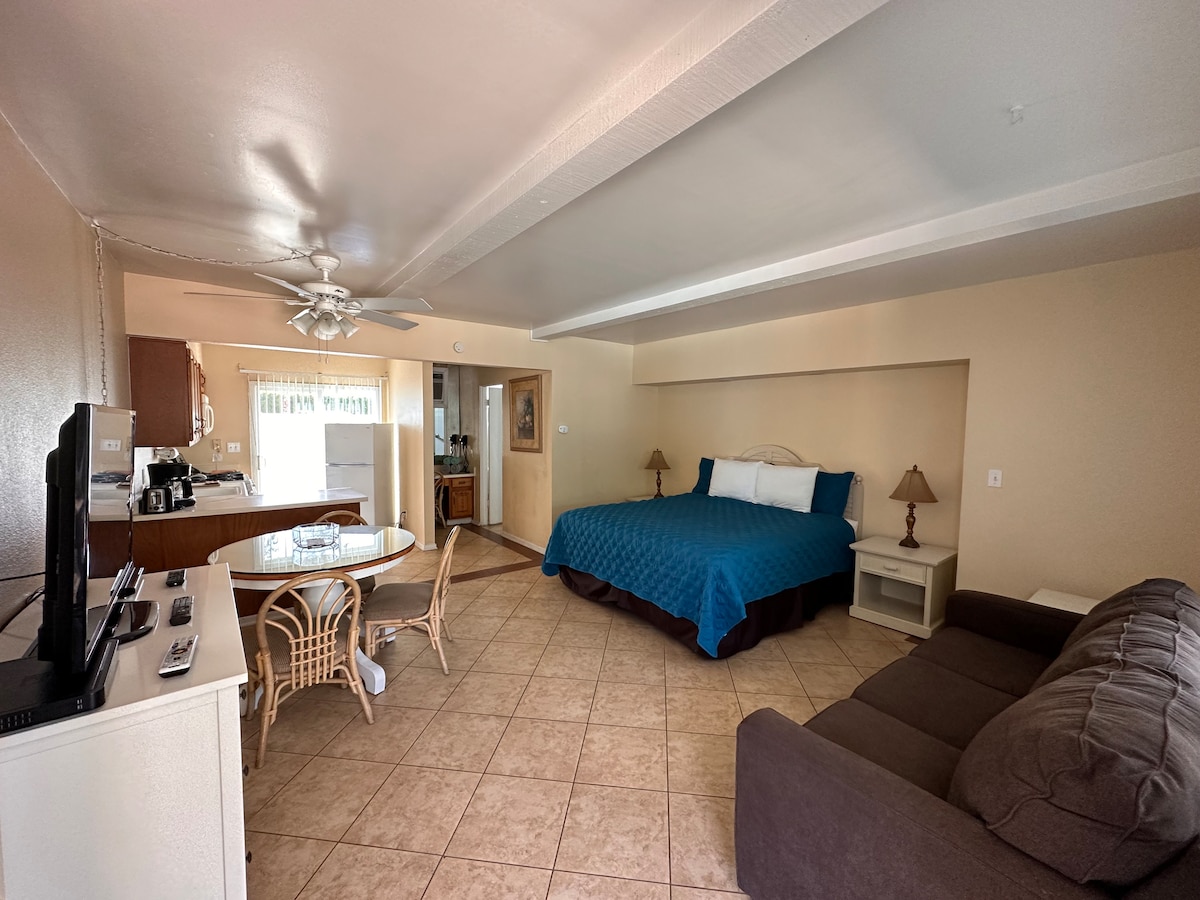 An open and airy studio layout features a spacious bed covered in a blue quilt. A couch and dining area can be seen, along with a kitchenette in the background. Natural light enters through a window, highlighting the tile flooring and neutral wall tones.