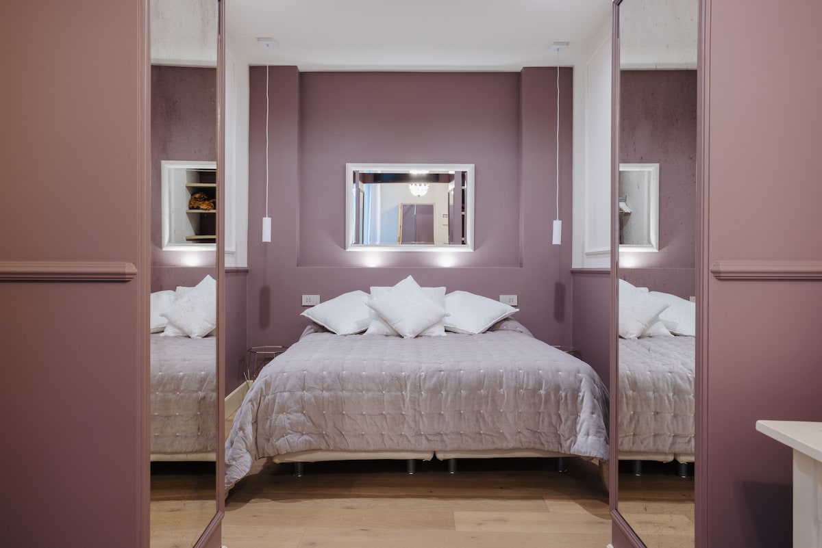 A spacious bedroom features a large bed adorned with neutral-hued bedding and decorative pillows. The walls are painted in soft lavender tones, complemented by mirrors that enhance the sense of space. Soft lighting is reflected by the central wall mirror, creating a calming ambiance.