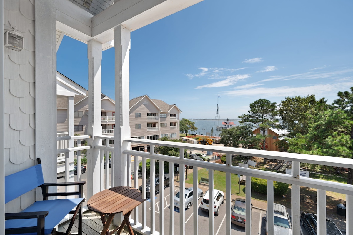 A balcony view showcases a clear sky above the waterfront, framed by white railings. Parking and grassy areas are visible below, along with nearby buildings. A small wooden table and a blue chair provide a cozy spot to enjoy the surroundings.