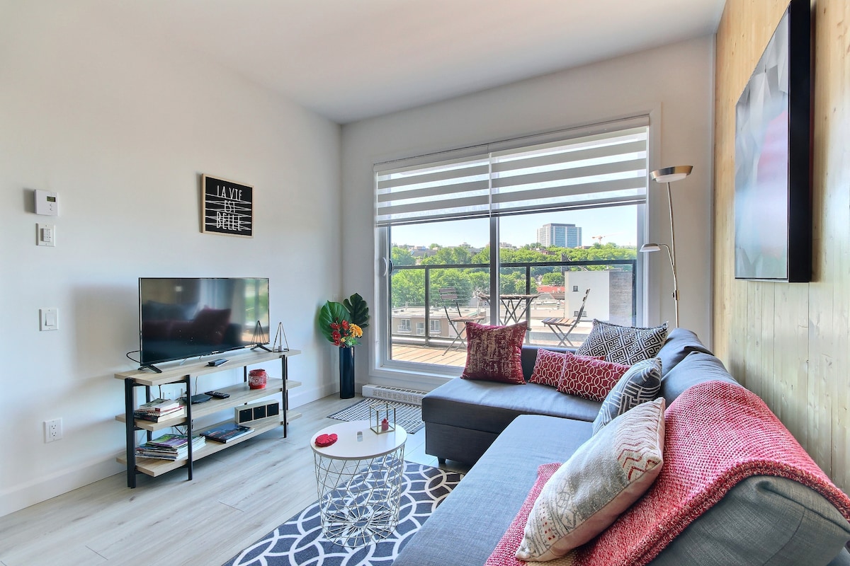 A modern living area features a gray sectional sofa adorned with decorative pillows in various patterns. A television is positioned on a stand with shelves below. Large windows allow natural light to fill the space, leading to a balcony with outdoor seating.