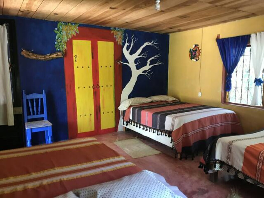 A colorful room featuring two spacious beds and a vibrant mural with a tree design. A blue chair is positioned in one corner, and large windows provide natural light, framed by white curtains. Decorative elements add a touch of charm to the space.