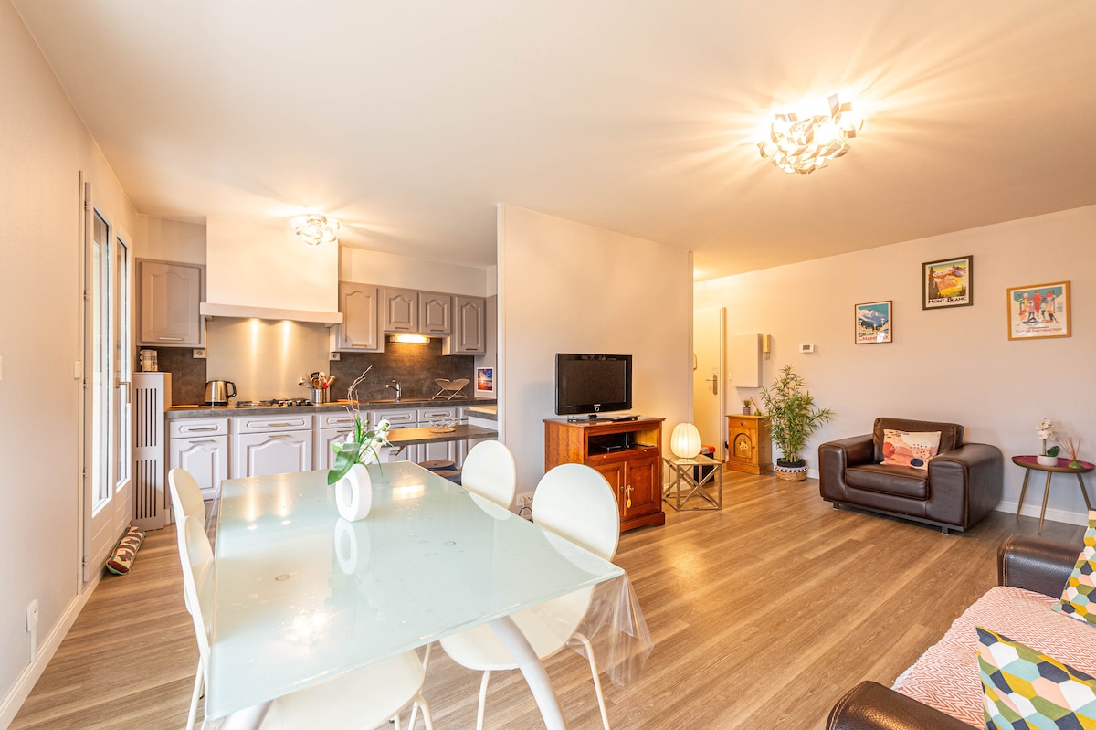 An open-plan living space features a well-equipped kitchen with modern appliances and gray cabinetry. A glass dining table is surrounded by white chairs. A comfortable seating area with dark furniture is visible, alongside a television, providing a functional and inviting environment.