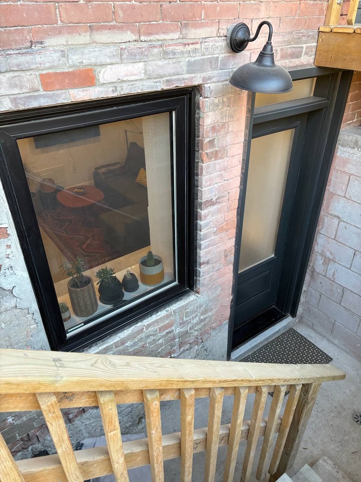An exterior view showcases the private entrance to the apartment. A dark door is framed by a large window, revealing glimpses of interior decor. Sturdy wooden stairs lead down from a balcony, and planters with greenery are positioned on a ledge beneath the window.