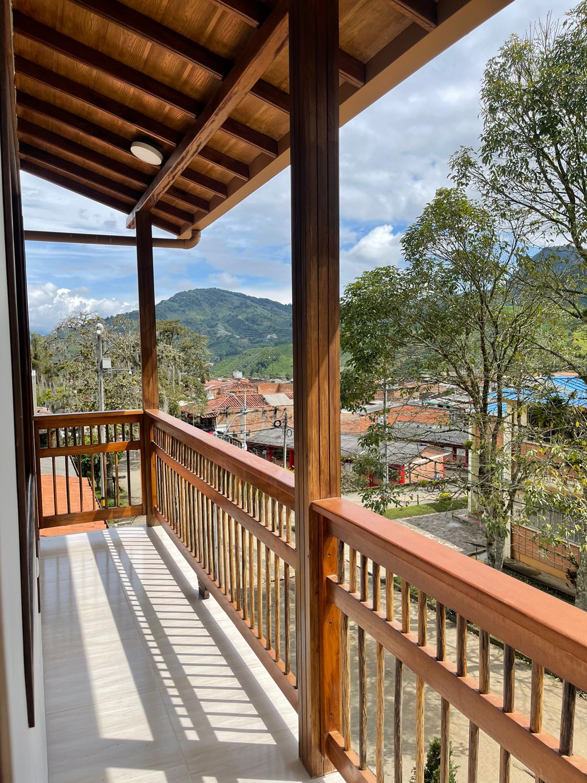 A spacious balcony is framed by wooden railings, offering views of surrounding mountains and nearby rooftops. Natural light illuminates the area, enhancing the wooden beams overhead and the tiled floor beneath. Lush trees can be seen in the distance, contributing to the serene setting.