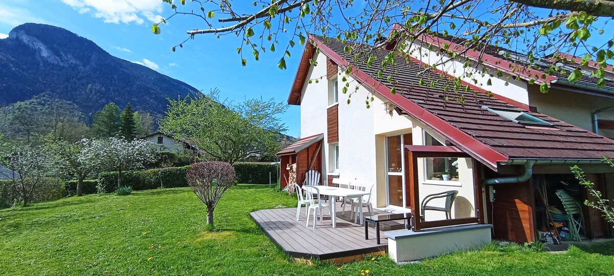 A two-story house is set against a backdrop of mountains, with a well-maintained lawn. A wooden deck features a table and chairs, providing an outdoor space for relaxation. Nearby, trees and shrubbery add greenery to the surroundings, enhancing the tranquil setting.