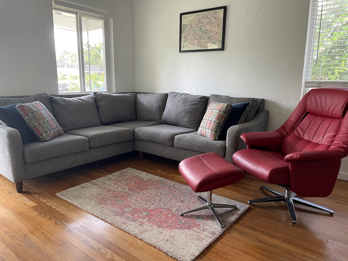 A modern living area features a spacious gray sectional sofa adorned with decorative pillows. A stylish red chair with an accompanying ottoman offers additional seating. Natural light enters through a nearby window, illuminating the warm hardwood floor and a patterned area rug.
