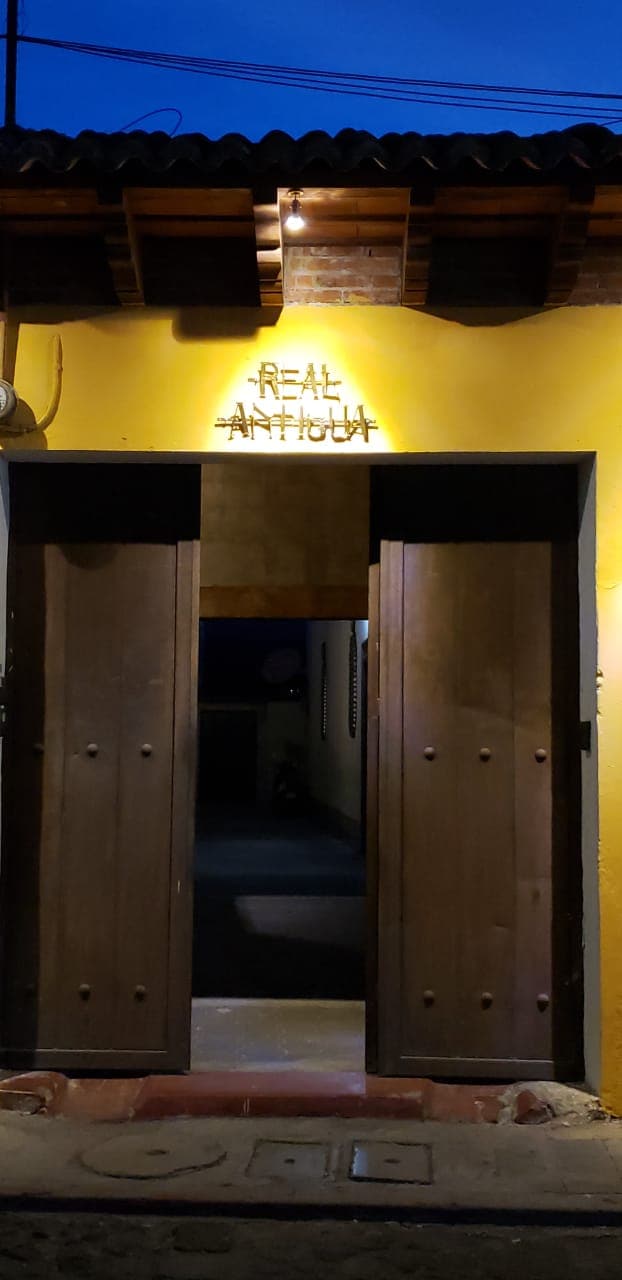 The entrance to the Hotel Boutique Real Antigua is featured, showcasing a warm yellow wall with the hotel name illuminated above. Two wooden doors provide access to a dimly lit corridor, inviting guests to explore the interior.