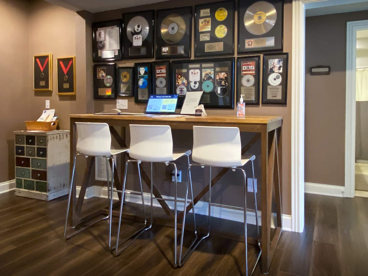 A modern bar area features three white stools positioned at a wooden counter. Walls are adorned with multiple framed music awards and gold records, reflecting the rich musical history of the space. The flooring is a dark wood, contributing to the contemporary feel.