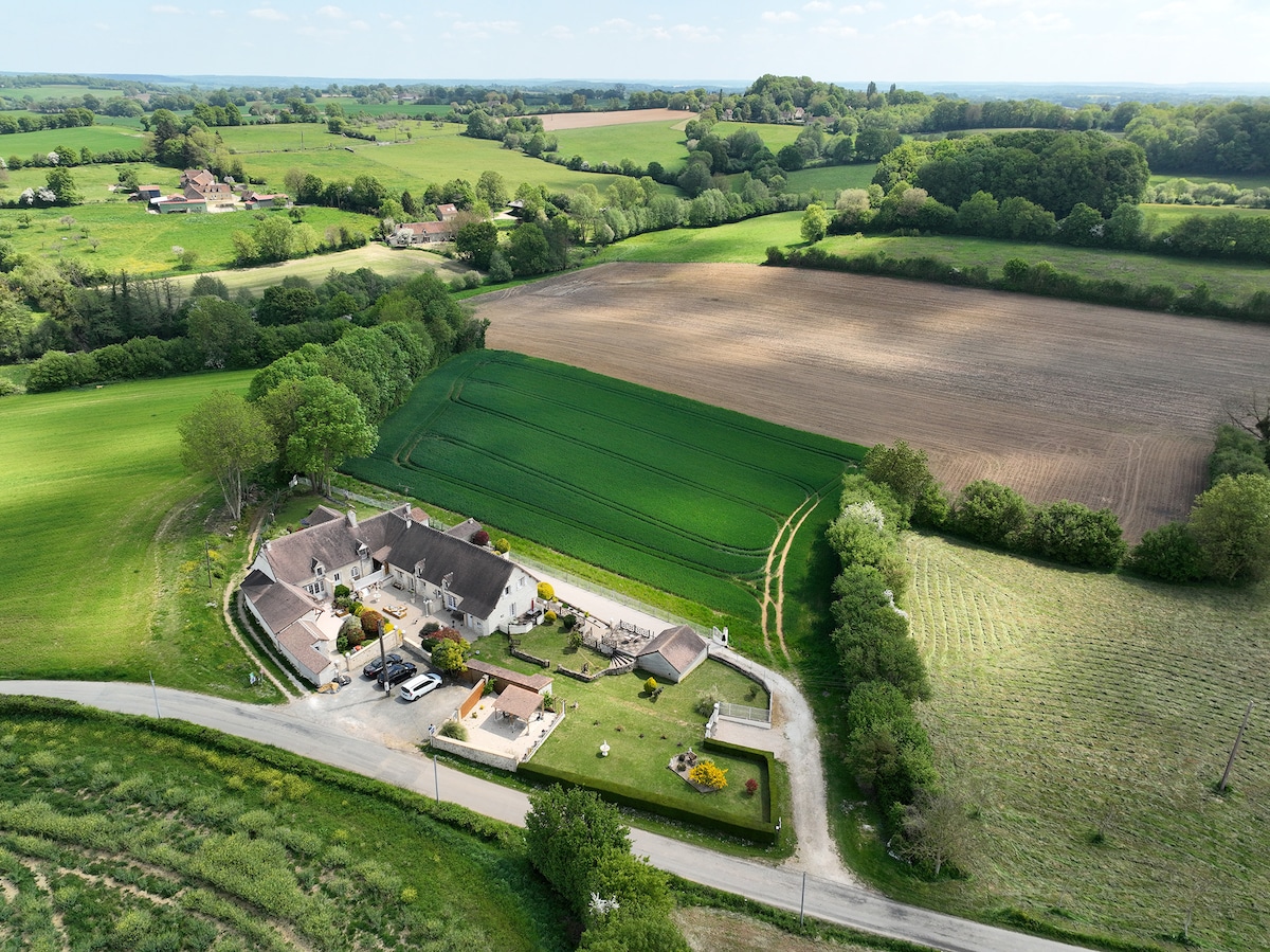An aerial view captures the charming gîte nestled in the countryside, surrounded by lush green fields and patches of cultivated land. The property is bordered by winding roads, with a peaceful rural landscape stretching into the distance.