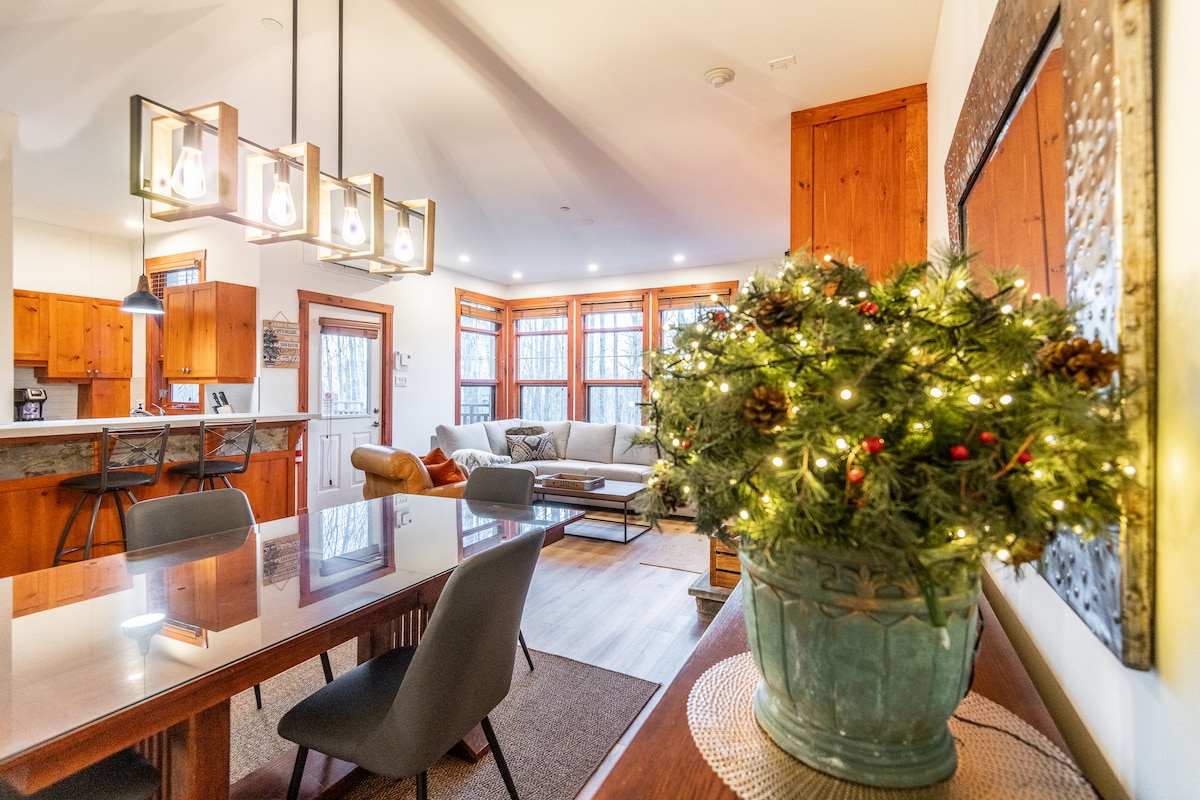 A dining area is seen with a table surrounded by chairs, positioned next to a living space featuring a light-colored couch and large windows. A decorative arrangement with pinecones and lights accents the table, creating a cozy ambiance.
