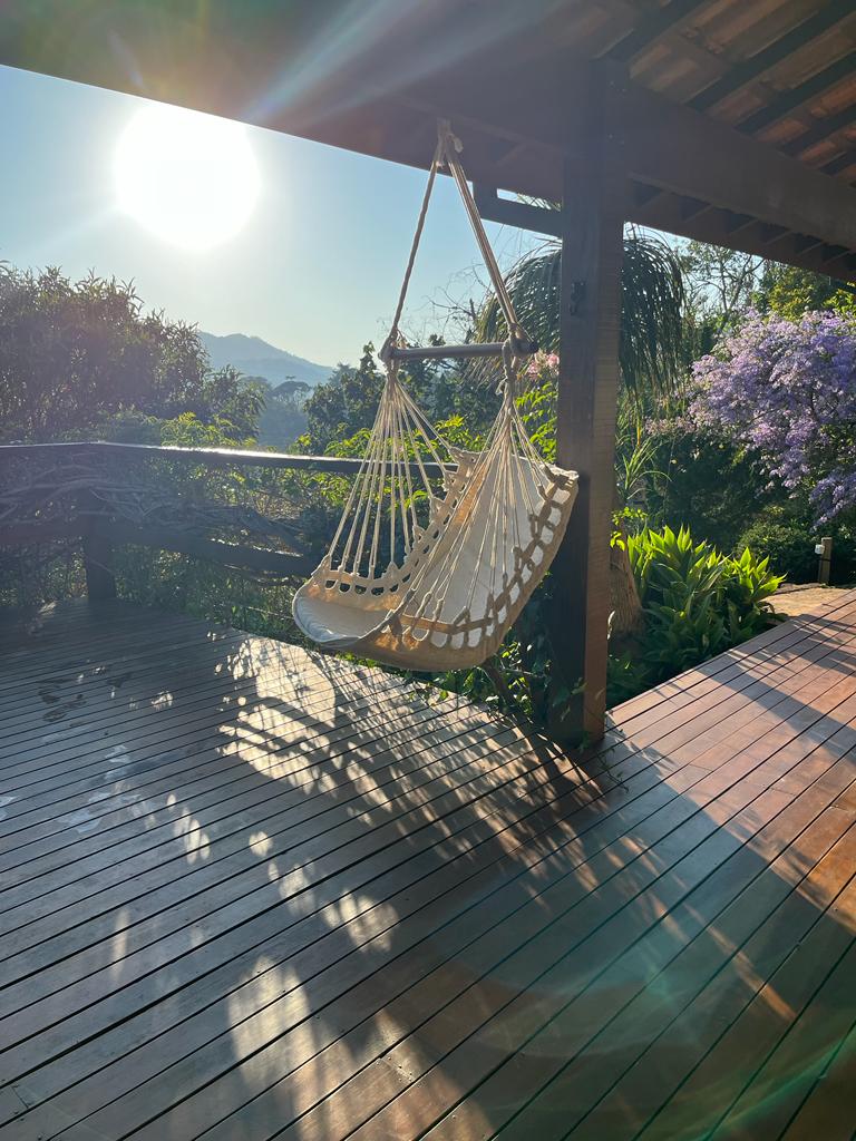 A hanging hammock sways gently under a covered deck, surrounded by lush greenery and vibrant blooming plants. The sun casts a warm glow, illuminating the wooden floor and offering a tranquil space for relaxation with mountain views in the distance.