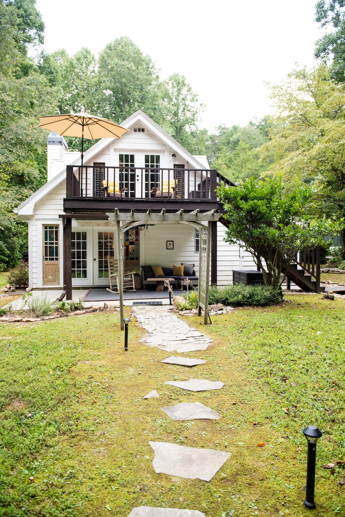 A charming farmhouse features a covered porch with outdoor seating and a large umbrella. A stone path leads to the entrance, surrounded by green grass and landscaping. The home is framed by trees, offering a serene setting in a peaceful environment.