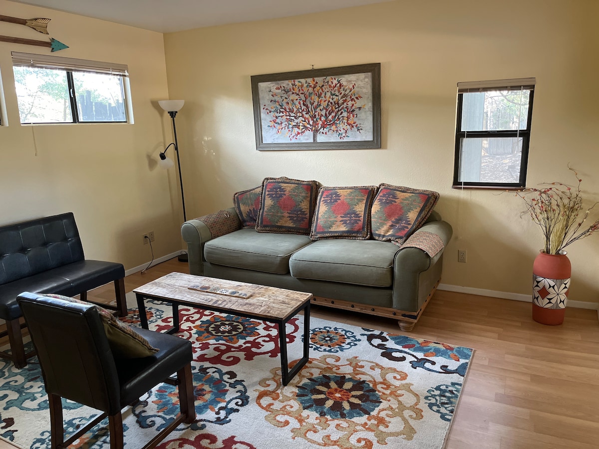 A cozy living area contains a plush sofa adorned with colorful patterned cushions. A wooden coffee table is positioned on a decorative area rug, while a tall lamp and wall art enhance the space. Natural light filters in through partially covered windows.