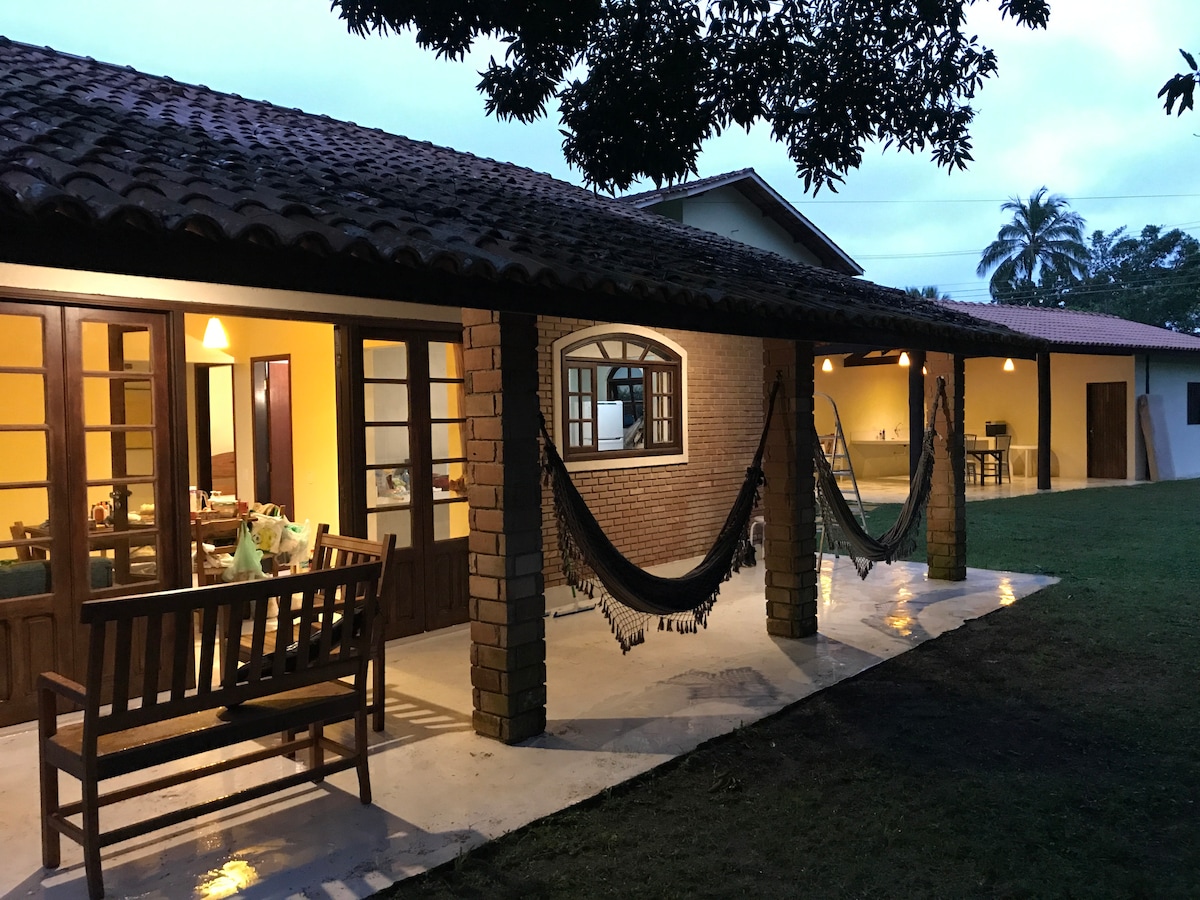 An outdoor area is depicted with a covered patio featuring wooden seating and two hammocks. The patio connects to the interior of the house, visible through large glass doors. A spacious garden is visible in the background, with a distant view of another structure.