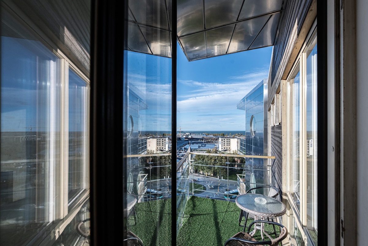 The image shows a glass-enclosed terrace offering expansive views of the surroundings. A green carpet covers the floor, while two round tables with chairs are positioned on the terrace, providing a comfortable space to enjoy the scenery.
