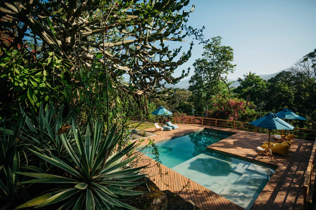 A serene pool area is surrounded by lush greenery and vibrant flora. Sun loungers are placed on a wooden deck, with umbrellas providing shade. The clear water of the pool reflects the sky, offering a refreshing atmosphere for relaxation.