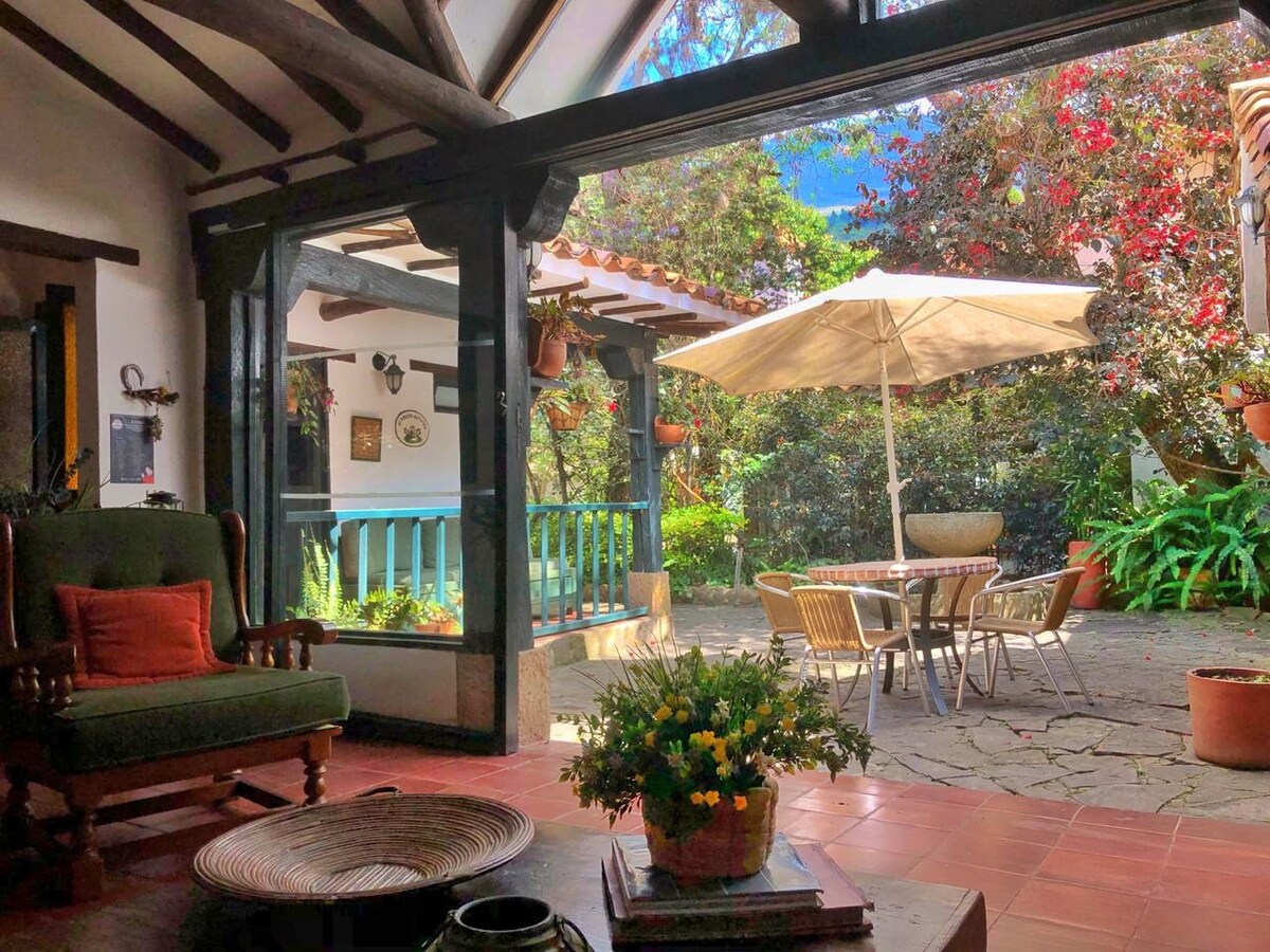 The interior living area features wooden beams and a comfortable green armchair. A door opens to a spacious patio, furnished with a table and dining chairs, shaded by a large parasol. Vibrant plants and flowers are visible in the background, enhancing the natural surroundings.
