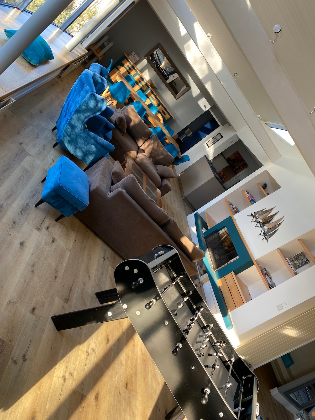 A spacious living area features a mix of comfortable seating arrangements, including brown sofas and blue armchairs. A foosball table is prominently located in the foreground, with a fireplace and shelving visible in the background, creating a welcoming atmosphere for gatherings.