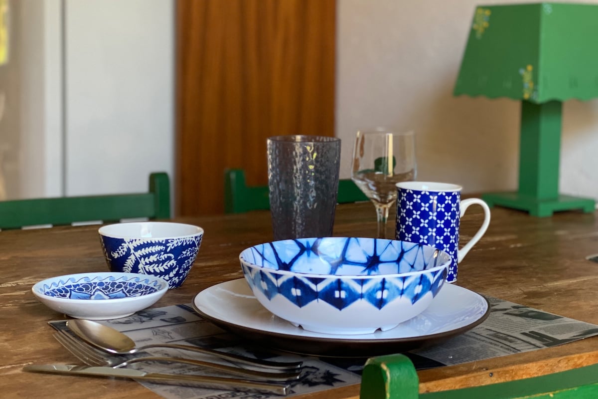 A dining table is set with an assortment of blue and white ceramic dishes, including bowls and plates. Glassware is positioned alongside silverware, and a green lamp provides subtle illumination. The natural wooden table adds warmth to the setting.