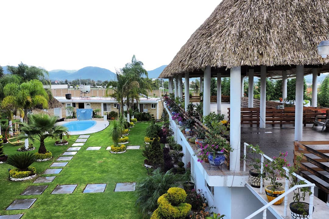 A well-maintained garden is adorned with diverse plants and flowers, leading to a glimmering swimming pool. A thatched palapa provides shaded seating, while stone pathways create an inviting connection between outdoor spaces. Lush green hills are visible in the background.