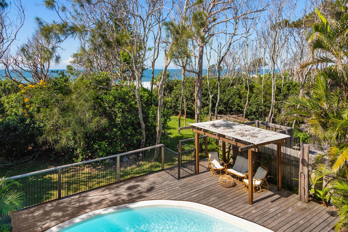 An inviting outdoor area features a saltwater pool surrounded by timber decking. A covered seating space with comfortable chairs sits nearby, framed by lush green trees. The ocean is visible beyond the greenery, enhancing the relaxed coastal setting.
