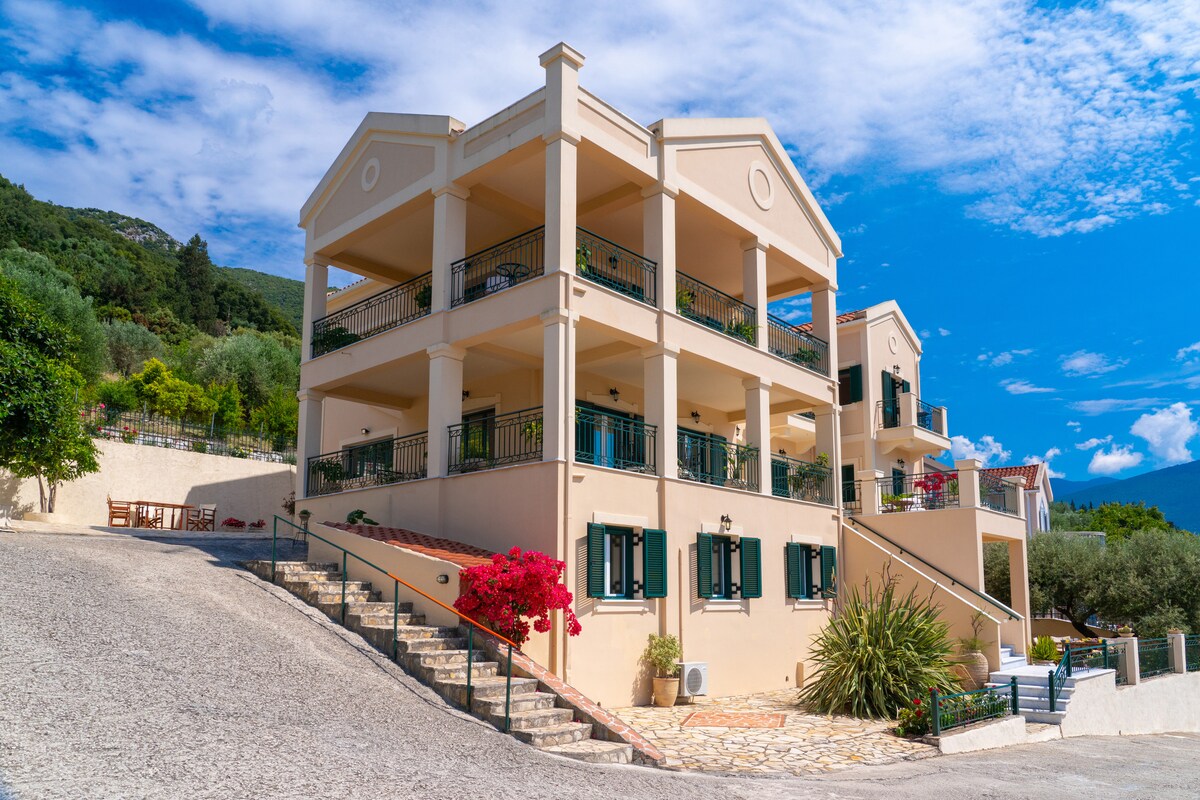 The two-story building is surrounded by lush greenery and features multiple balconies with black railings. The facade is painted in soft tones, complemented by dark green shutters. Colorful flowering plants add vibrancy to the landscaped path leading to the entrance.