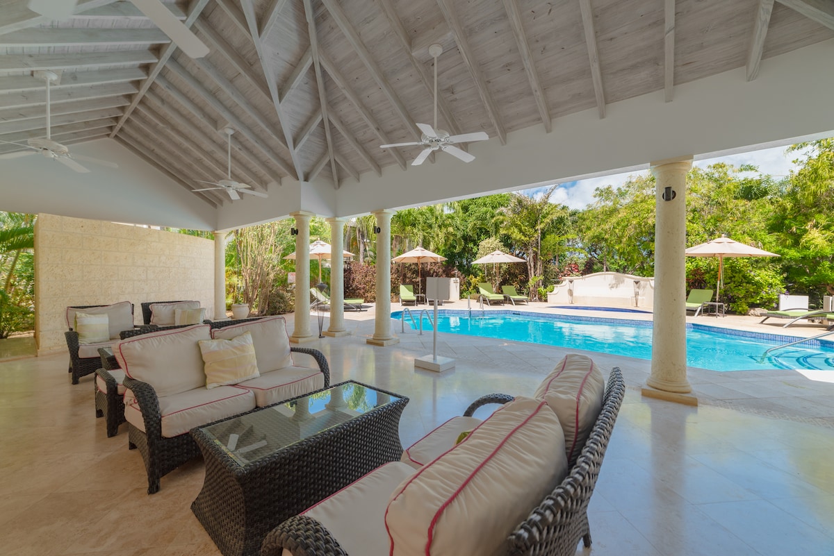 A covered patio area features comfortable seating with plush cushions surrounding a glass-top table. Ceiling fans provide a gentle breeze, and a sparkling pool is visible in the background, complemented by shaded lounge chairs and tropical greenery.