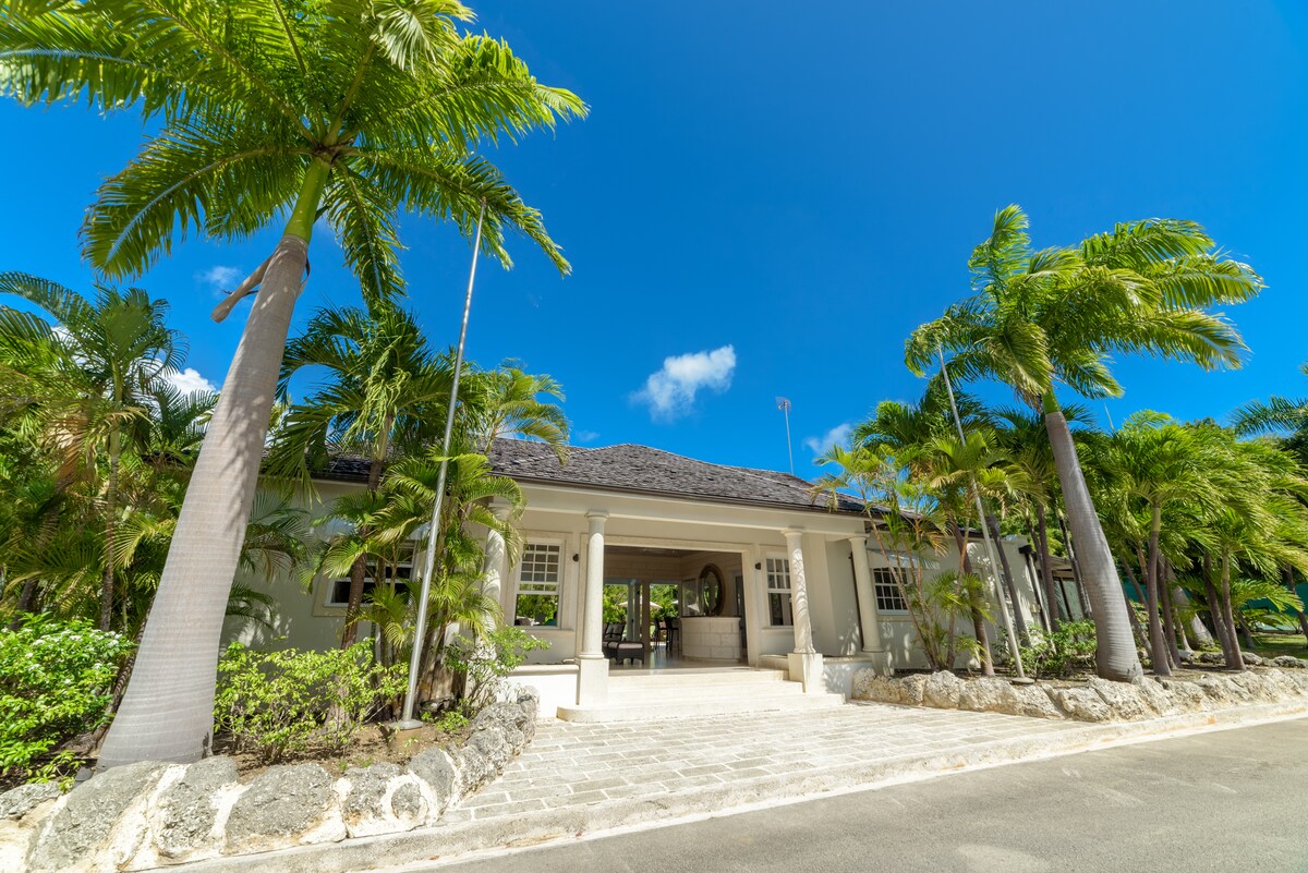 The entrance of the villa is framed by tall palm trees and lush greenery, showcasing a wide, stone pathway that leads to the main door. A smoothly tiled area extends from the front entrance, complemented by the clear blue sky above.