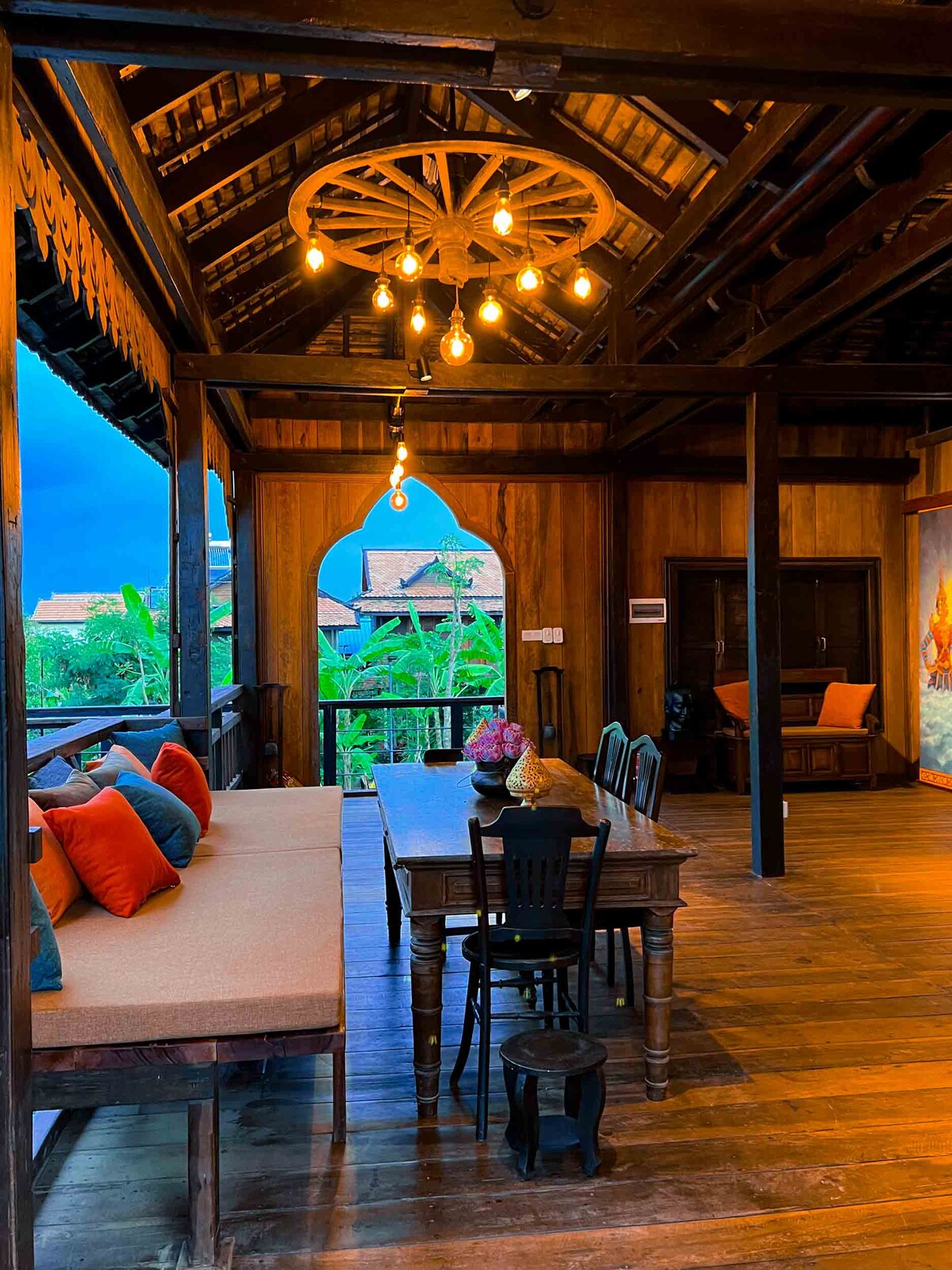 An open living space features high wooden ceilings with a rustic chandelier. A long dining table is accompanied by black chairs, while a cozy seating area with vibrant cushions invites relaxation. Large windows showcase the surrounding greenery, enhancing the room's natural ambiance.